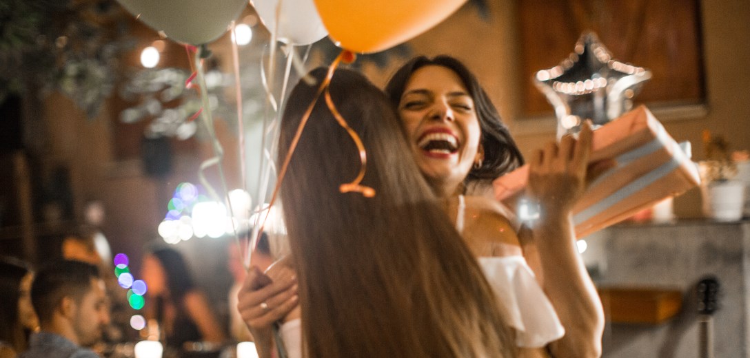 Two women are hugging. One of the two women holds a present and balloons in her hands and laughs happily.