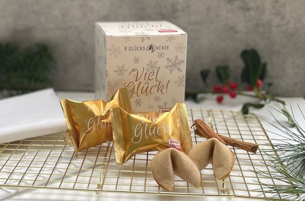 Glückskekse Faltschachtel und in Goldfolie mit Viel Glück-Aufschrift und zwei ausgepackte Kekse auf einem goldenen Rost mit Zimtstange und Tannengrün im Hintergrund