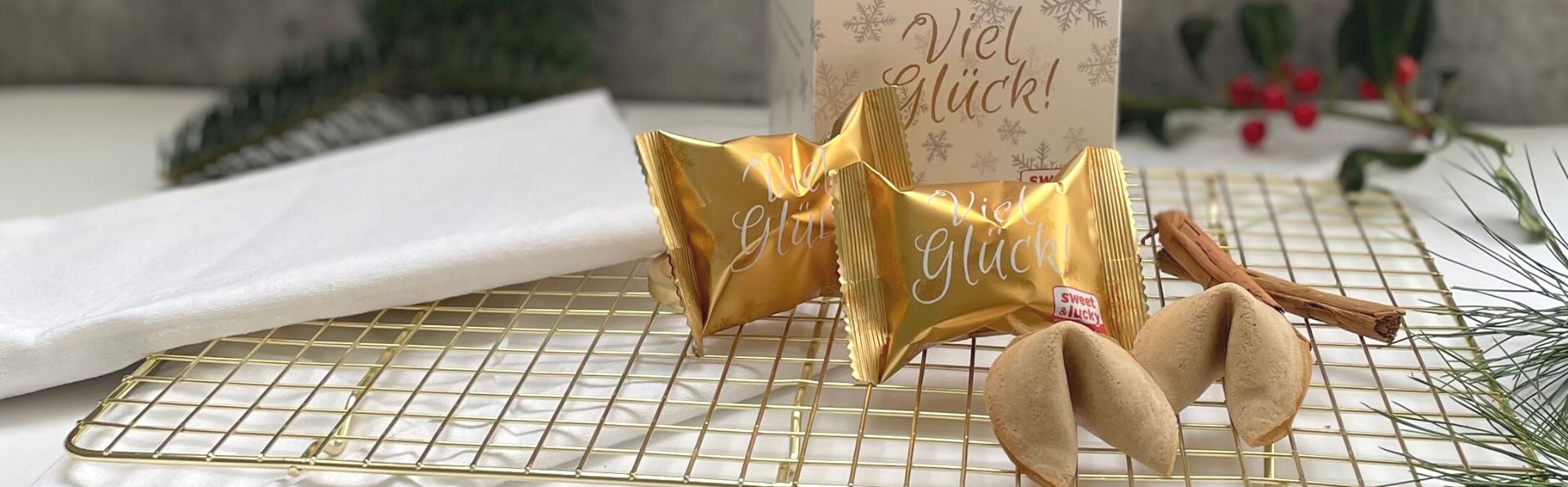 Glückskekse Faltschachtel und in Goldfolie mit Viel Glück-Aufschrift und zwei ausgepackte Kekse auf einem goldenen Rost mit Zimtstange und Tannengrün im Hintergrund
