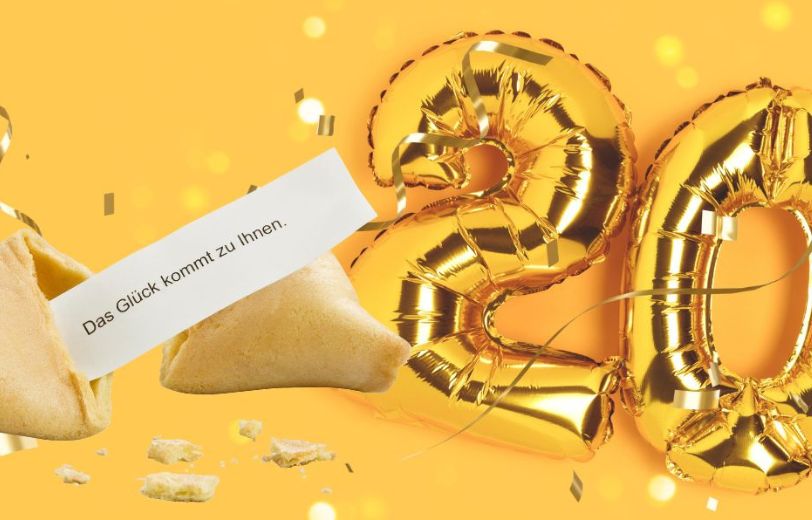20 als goldene Folienballons vor gelbem Hintergrund mit goldenen Luftschlangen und Konfetti mit einem offenen Glückskeks