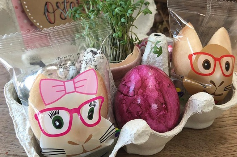 Ein Eierkarton ist mit zwei Glückskeksen, einem Osterei sowie Blumen geschmückt und wird so zum perfekten Oster Mitbringsel.